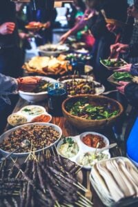 A festive potluck table with people dishing out beans, salads, hummus and more