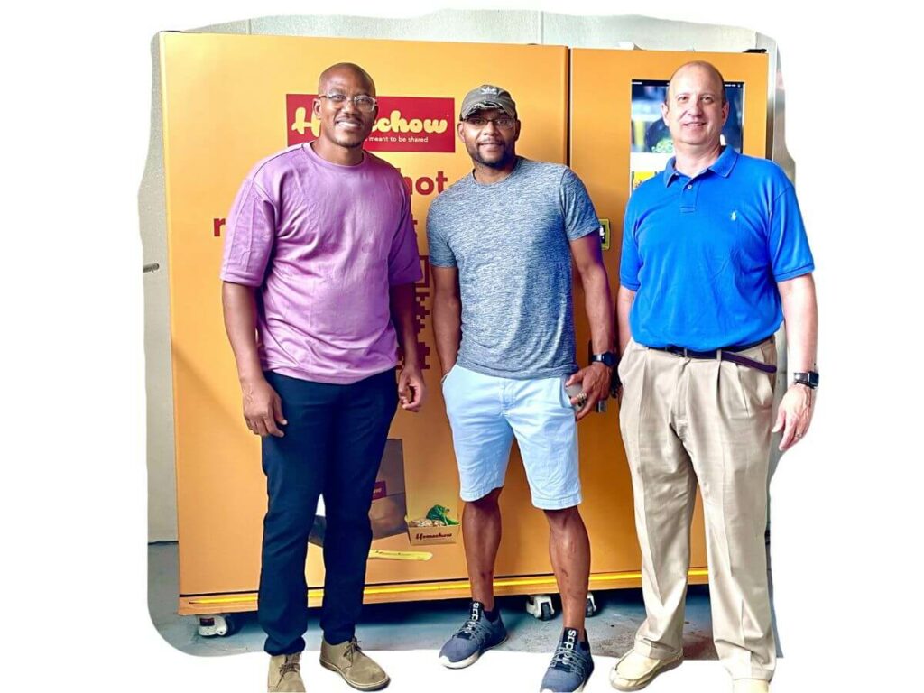 Three men standing in front of a Homechow kiosk smiling