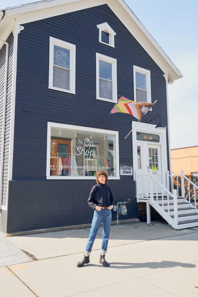 Woman in jeans and a black shirt smiling in front of The Op Shop in Rochester, NY.